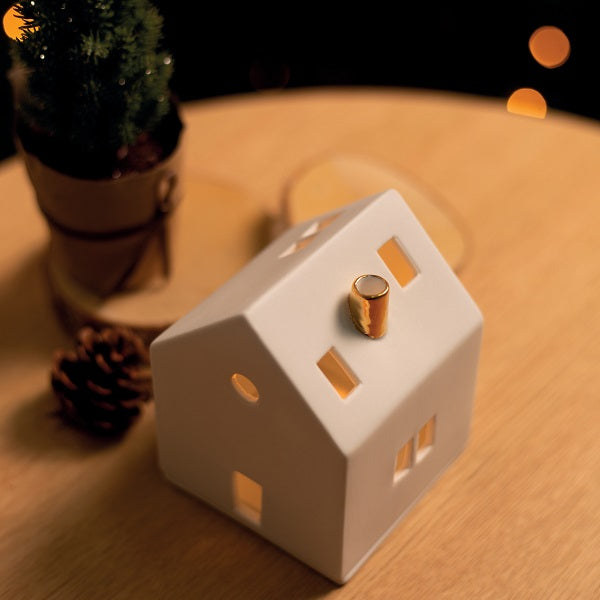 Photophore en porcelaine blanche en forme de maison avec une cheminée dorée, sur une table.