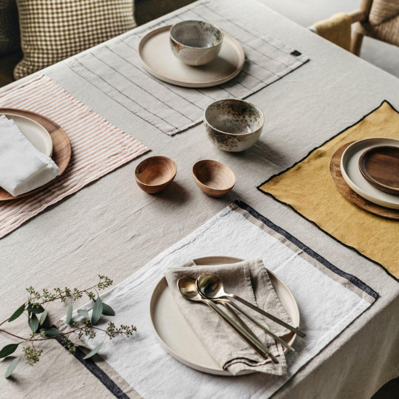 Table dressée avec une nappe unie en lin avec des sets de tables différents et de la vaisselle. 
