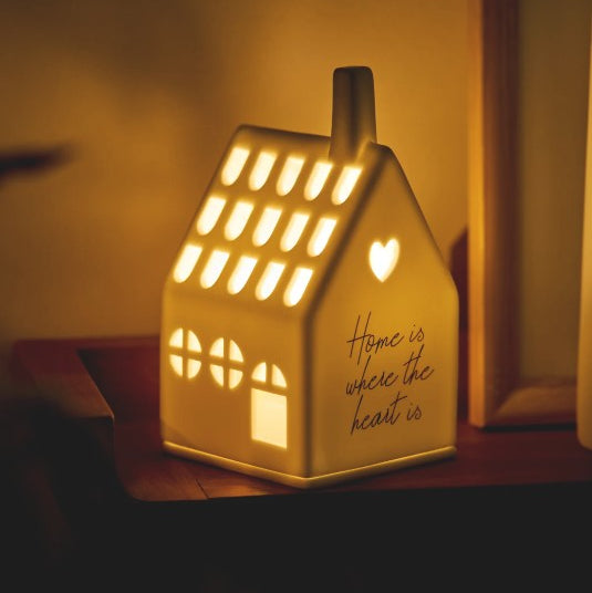 Photophore en porcelaine blanche ajourée en forme de maison, avec une citation en façade, allumé.