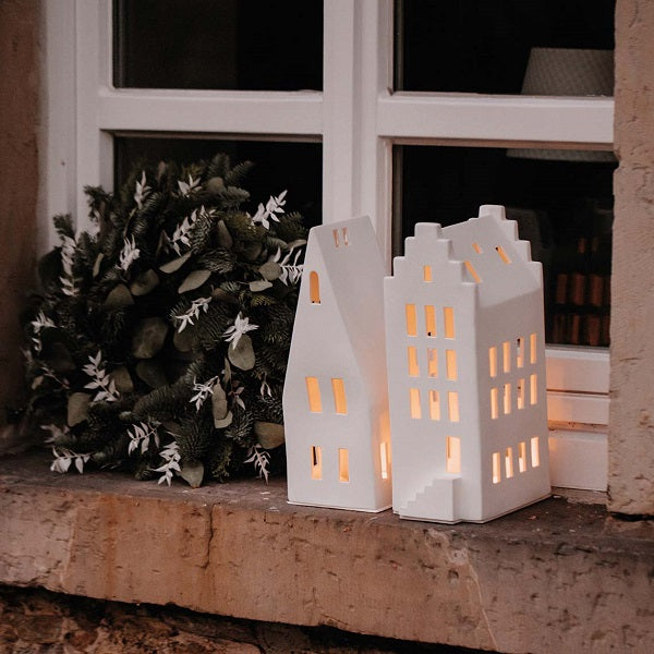 Photophore en porcelaine blanche en forme de maison avec un grand toit pentu, sur un rebord de fenêtre.